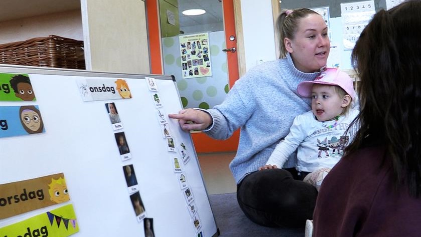Förskolepedagog sitter med ett barn i knä och pekar på en tavla med små bilder på 