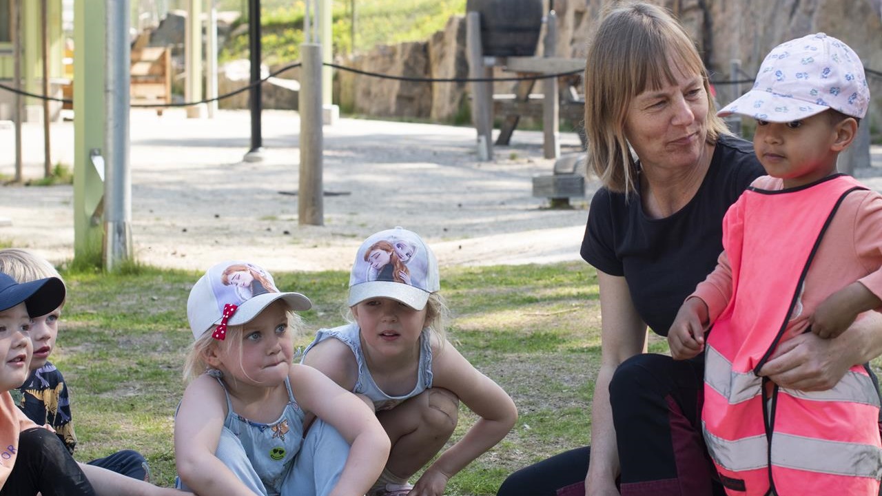 En förskolepedagog står på knä vid ett barn och håller om och tittar på det. Några barn sitter bredvid 